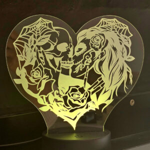 Una lámpara de acrílico en forma de corazón muestra dos calaveras, una masculina y otra femenina, besándose, rodeadas de rosas y telarañas, creando una atmósfera gótica romántica.