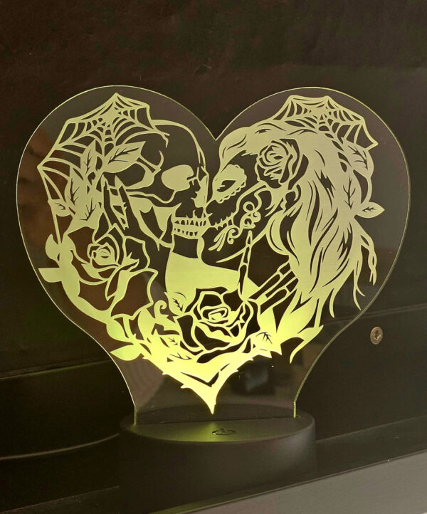 Una lámpara de acrílico en forma de corazón muestra dos calaveras, una masculina y otra femenina, besándose, rodeadas de rosas y telarañas, creando una atmósfera gótica romántica.